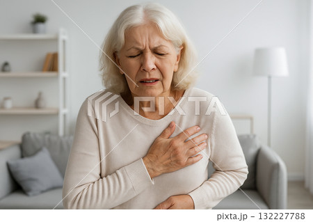 Senior woman clutching chest with painful expression in modern living room. Concept of heart attack, panic attack, chest pain, cardiovascular emergency, senior health issue, elderly woman in distress Senior woman clutching chest with painful expression in modern living room. Concept of heart attack, panic attack, chest pain, cardiovascular emergency, senior health issue, elderly woman in distress 132227788