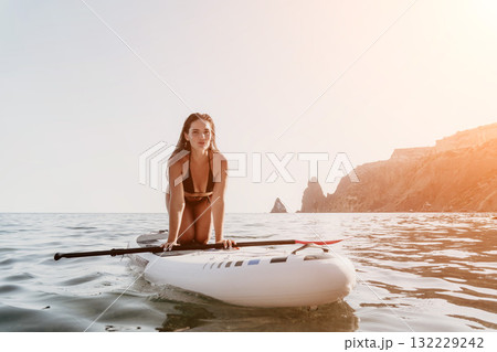 Woman Paddleboard Ocean - A woman in a black bikini kneels on a white paddleboard in the ocean, with rocky cliffs in the background. Woman Paddleboard Ocean - A woman in a black bikini kneels on a white paddleboard in the ocean, with rocky cliffs in the background. 132229242