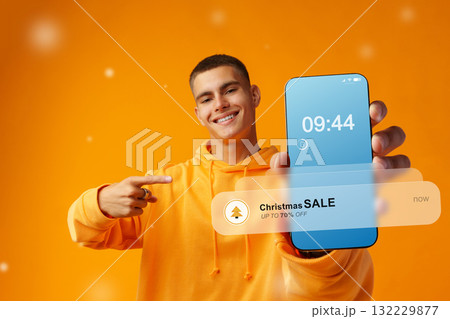 Cheerful young man showing smartphone with Christmas sale notification at morning Cheerful young man showing smartphone with Christmas sale notification at morning 132229877