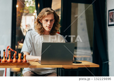 Young man working on laptop at a cafe with chess pieces nearby during the day Young man working on laptop at a cafe with chess pieces nearby during the day 132229896