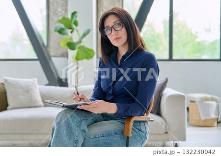 Portrait of serious business woman with notebook sitting in chair in office Portrait of serious business woman with notebook sitting in chair in office 132230042