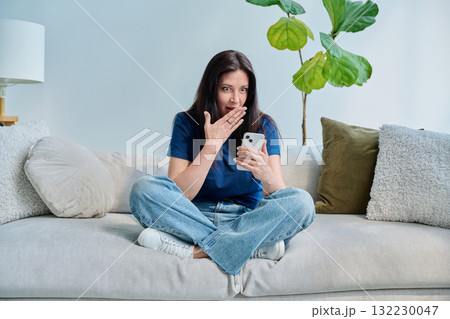 Young surprised woman sitting at home on the couch using smartphone Young surprised woman sitting at home on the couch using smartphone 132230047