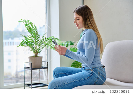 Young woman sitting at home in chair using smartphone 132230051