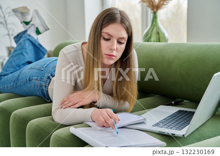 Young female student lying at home on couch with laptop, notebook, textbook 132230169