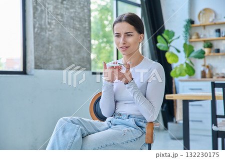 Teenage girl sitting at home in chair with smartphone, recording an audio message Teenage girl sitting at home in chair with smartphone, recording an audio message 132230175