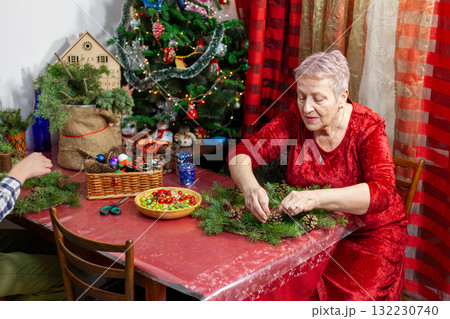 Senior old woman craft. Grandmother make Christmas wreath from spruce branches. Home decoration in December Senior old woman craft. Grandmother make Christmas wreath from spruce branches. Home decoration in December 132230740