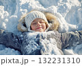 A joyful child lies in soft, fresh snow with arms spread wide, creating snow angels under clear blue skies. The winter scene showcases bright sunlight reflecting off the snow 132231310