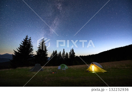 Several tents on grassy clearing under breathtaking, star-filled sky with Milky Way prominently visible. Pine trees and distant mountains frame scene, creating awe-inspiring camping experience. 132232936
