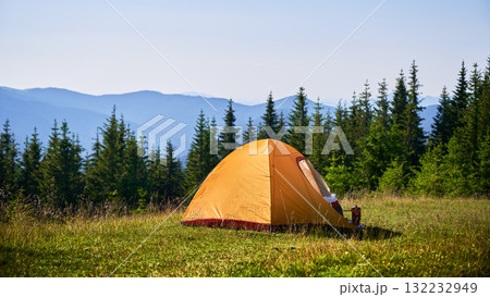 Bright orange tourist tent on grassy hillside, surrounded by lush green pine trees. Breathtaking view of distant blue mountains under clear sky, creating idyllic and serene camping spot. Bright orange tourist tent on grassy hillside, surrounded by lush green pine trees. Breathtaking view of distant blue mountains under clear sky, creating idyllic and serene camping spot. 132232949