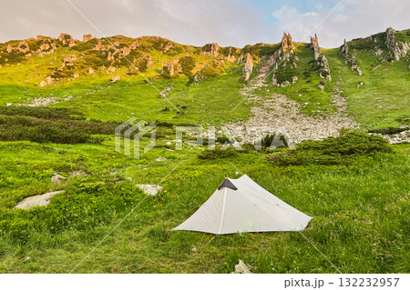 Lone white tourist tent in lush green valley surrounded by towering hills and rocky cliffs. Sky above shows hint of sunrise colors, creating serene camping scene. Carpathian mountains, Ukraine. 132232957