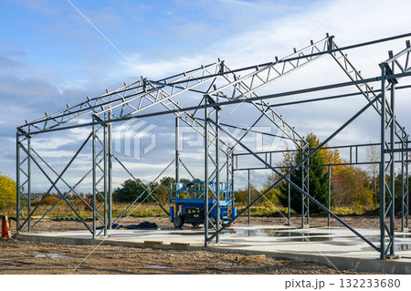 Steel frame of industrial warehouse under construction with scaffolding and blue lift on a sunny day Steel frame of industrial warehouse under construction with scaffolding and blue lift on a sunny day 132233680