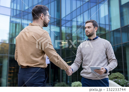 Two professional businessmen shaking hands in an urban setting, solidifying their partnership and reaching a positive agreement outside a modern office building 132233744