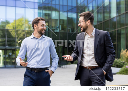 Two smiling adult businessmen walking and having a conversation outside a modern corporate office building, discussing business strategy or partnership Two smiling adult businessmen walking and having a conversation outside a modern corporate office building, discussing business strategy or partnership 132233817