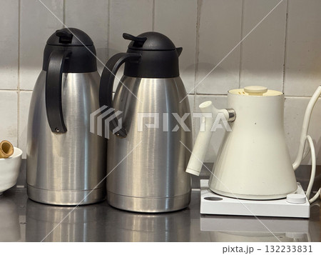 Metal thermoses and electric kettle on kitchen counter. Functionality, warmth and modern beverage preparation in cafe interior. Metal thermoses and electric kettle on kitchen counter. Functionality, warmth and modern beverage preparation in cafe interior. 132233831