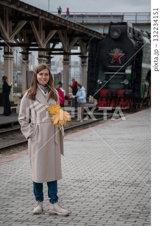 Woman with autumn leaves on her shoulder stands near an old steam locomotive at a railway station in Moscow 132234151