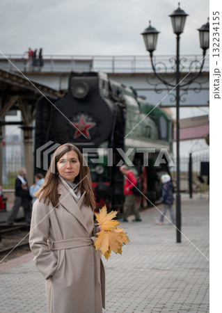 Woman with autumn leaves on her shoulder stands near an old steam locomotive at a railway station in Moscow Woman with autumn leaves on her shoulder stands near an old steam locomotive at a railway station in Moscow 132234155