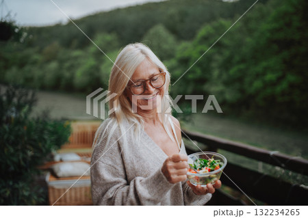 Senior woman standing terrace, eating healthy dinner outdoors. 132234265