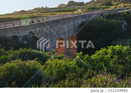 Historic stone bridge along Victoria Lines in Malta, glowing arches at sunset, green valley and shrubs in foreground Historic stone bridge along Victoria Lines in Malta, glowing arches at sunset, green valley and shrubs in foreground 132234460