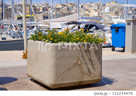 Square stone planter filled with greenery and small blooms on waterfront walkway, boats and harbor details in background, bright summer light 132234479