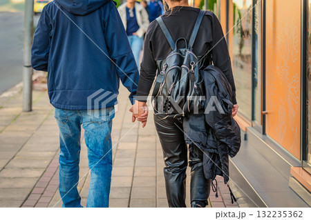 Young couple walks hand in hand on sidewalk, dressed in casual streetwear and leather. Concept of love, relationship, urban romance, casual urban lifestyle Young couple walks hand in hand on sidewalk, dressed in casual streetwear and leather. Concept of love, relationship, urban romance, casual urban lifestyle 132235362