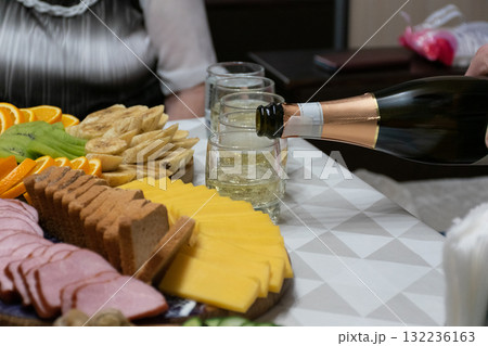Close-up of a festive table with sliced cheese, sausage, fruits, and biscuits as champagne is poured into a glass. Celebration scene filled with warmth, delicious food, and joyful atmosphere 132236163