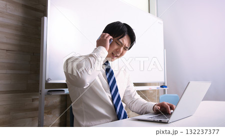オフィスでパソコンを見ながらスマホで通話する若いビジネスマン 132237377
