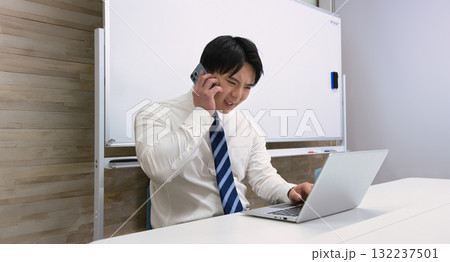 オフィスでパソコンを見ながらスマホで通話する若いビジネスマン オフィスでパソコンを見ながらスマホで通話する若いビジネスマン 132237501