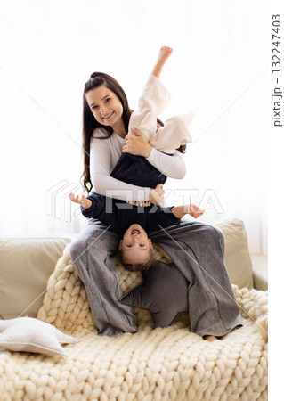 A mother holds her child upside down on a cozy couch, both smiling and enjoying a playful moment together. A mother holds her child upside down on a cozy couch, both smiling and enjoying a playful moment together. 132247403