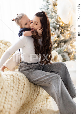 A young child kisses their mother's cheek, creating a tender moment in front of a Christmas tree. A young child kisses their mother's cheek, creating a tender moment in front of a Christmas tree. 132247415