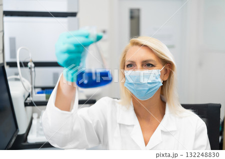 Female scientist wearing mask and lab coat, examining blue chemical solution in glass flask in laboratory. Female scientist wearing mask and lab coat, examining blue chemical solution in glass flask in laboratory. 132248830