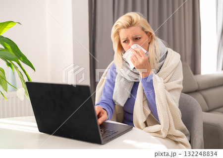 Middle aged blonde woman feeling unwell while working on her laptop from home. Concept of online work, health, and remote professional life during illness. 132248834