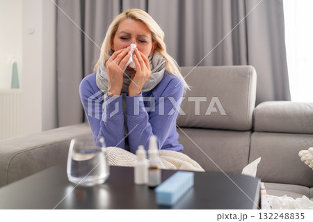 Sick middle aged woman wearing scarf sits on sofa, blowing her nose with tissue. Medicines and a glass of water on the table 132248835
