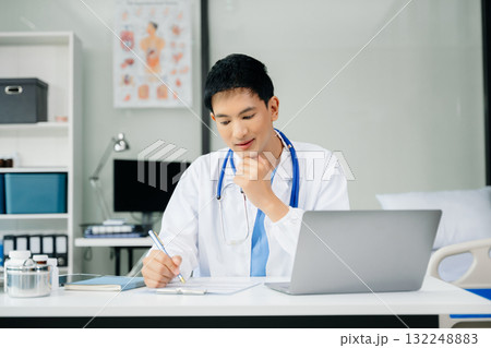 Confident young  male doctor in white medical uniform sit at desk working on computer. Smiling use laptop write in medical journal 132248883