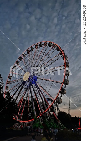 A giant Ferris wheel lit up with glowing orange lights at dusk in a popular Saint Petersburg amusement park, with visitors walking below. A giant Ferris wheel lit up with glowing orange lights at dusk in a popular Saint Petersburg amusement park, with visitors walking below. 132249009