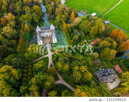 Aerial view of historic castle symmetrical twin towers and central hall framed by forest, moats, and tree lined path, with open fields and residential zone beyond under warm sunrise sky. 132249818