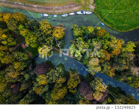Aerial view of lush forest narrow green tinted waterway with docked boats, small footbridge connects wooded banks, vibrant foliage and grassy clearing evoke serene natural harmony. 132249825