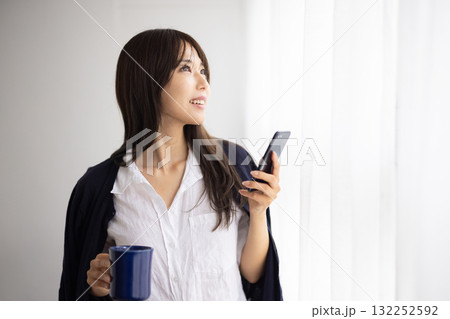 窓辺でマグカップを持ちながらスマートフォンを操作する笑顔の女性 窓辺でマグカップを持ちながらスマートフォンを操作する笑顔の女性 132252592
