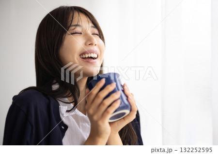 窓辺でマグカップを持ち微笑む若い女性 窓辺でマグカップを持ち微笑む若い女性 132252595