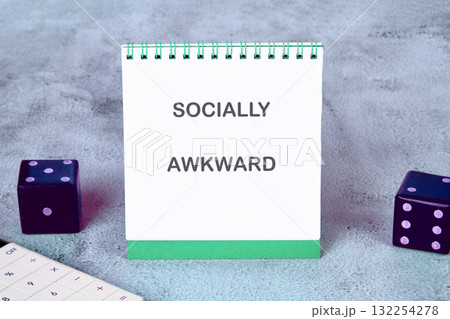 A notepad with the words socially awkward stands prominently on a soft gray surface beside two colorful dice. This scene invites reflections on personal experiences and humorous situations 132254278