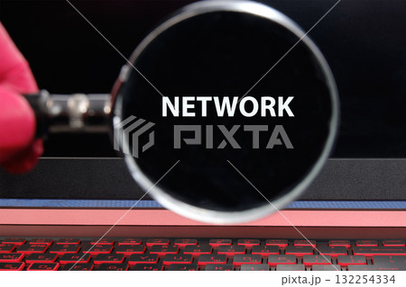 A hand holds a magnifying glass focused on the word NETWORK displayed on a dark laptop screen, symbolizing the exploration of digital connections and technology 132254334