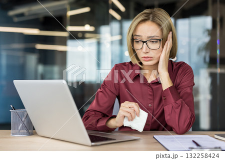 Businesswoman feeling upset and overwhelmed, holding a tissue while looking at her laptop screen in the office, conveying feelings of sadness, bad news, and burnout 132258424