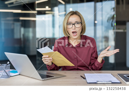 Businesswoman looking shocked and stressed while opening an official envelope with important documents, sitting at a desk in a modern office, reacting to unexpected problems or a big surprise Businesswoman looking shocked and stressed while opening an official envelope with important documents, sitting at a desk in a modern office, reacting to unexpected problems or a big surprise 132258438