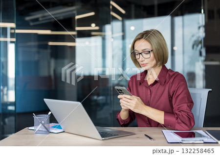 Businesswoman with glasses managing communication and tasks on her smartphone while sitting at a wooden desk with a laptop and documents , in a contemporary corporate office environment Businesswoman with glasses managing communication and tasks on her smartphone while sitting at a wooden desk with a laptop and documents , in a contemporary corporate office environment 132258465