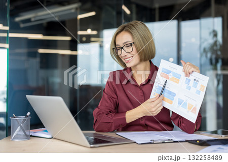 Businesswoman giving a financial presentation during a video call, confidently pointing at a chart while working remotely from a modern office using her laptop Businesswoman giving a financial presentation during a video call, confidently pointing at a chart while working remotely from a modern office using her laptop 132258489