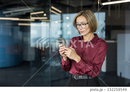 Businesswoman with glasses standing in a contemporary office space, focused on her smartphone, engaging in digital communication and managing her professional tasks 132258549