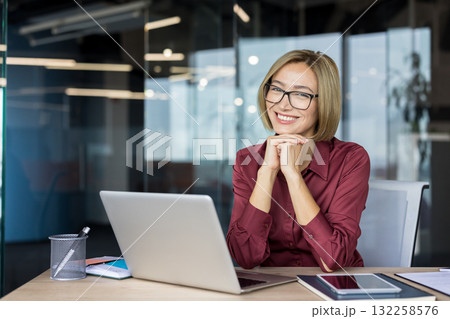 Professional businesswoman smiling confidently, resting chin on hands at desk with laptop in modern office, glasses, relaxed and focused portrait conveying success and leadership Professional businesswoman smiling confidently, resting chin on hands at desk with laptop in modern office, glasses, relaxed and focused portrait conveying success and leadership 132258576