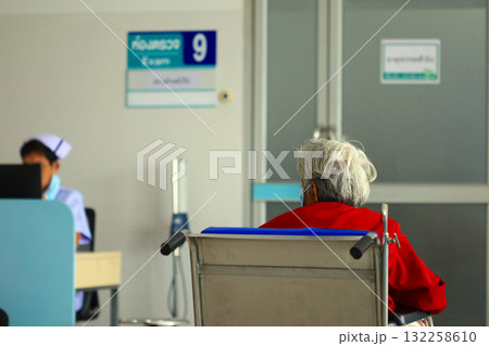 An elderly woman with gray hair in a red dress sits in a wheelchair and waits in a hospital examination room with a sign in Thai. A nurse stands behind her. An elderly woman with gray hair in a red dress sits in a wheelchair and waits in a hospital examination room with a sign in Thai. A nurse stands behind her. 132258610