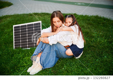 Mother and daughter sit together on grass, embracing in warm moment with solar panel beside in the evening. Concept of renewable energy and family, highlighting commitment to sustainable future. 132260827