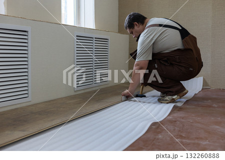 A worker, kneeling, carefully lays laminate flooring boards over an insulated underlayment, transforming a room during a home improvement and renovation project. Laminate flooring installation. 132260888