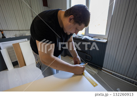Man carefully measuring and marking a kitchen cabinet panel with ruler and pencil, preparing to assemble flat-pack furniture during home renovation or diy installation project. 132260913